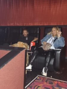 Two adults sit in a movie theater, one with a drink, one with a snack.